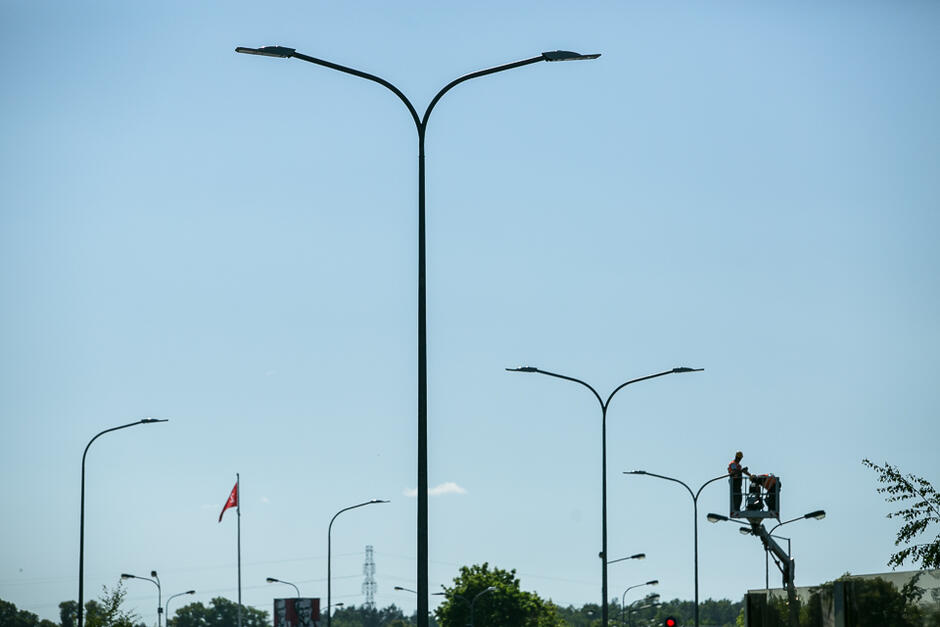 Na zdjęciu widoczni są dwaj pracownicy na podnośniku, którzy dokonują wymiany opraw oświetleniowych na jednym ze słupów latarni ulicznych.